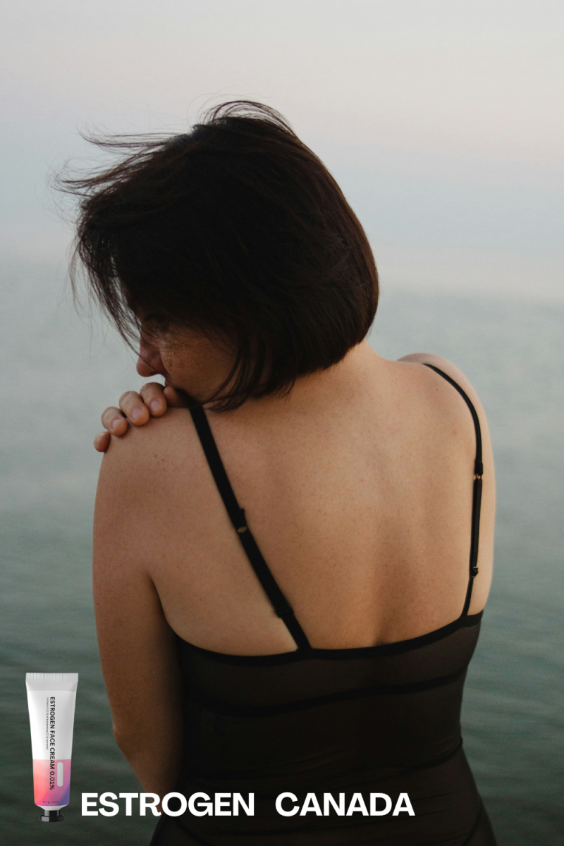 Woman with short dark hair in black top looks at sea, back to viewer.