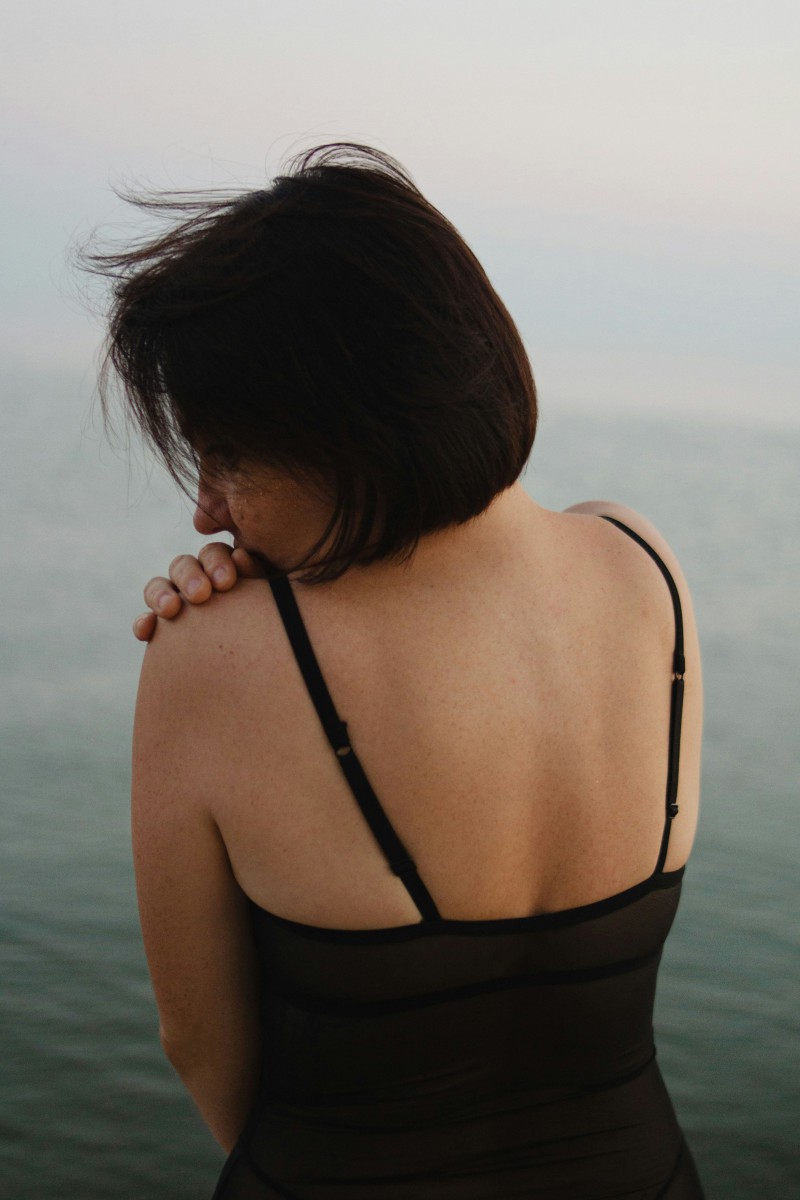 Woman with short dark hair in black top looks at sea, back to viewer.